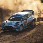 A white and blue Ford rally car speeds along a rugged dirt track, kicking up a massive cloud of thick dust behind it