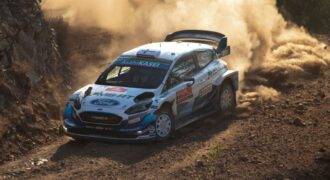 A white and blue Ford rally car speeds along a rugged dirt track, kicking up a massive cloud of thick dust behind it