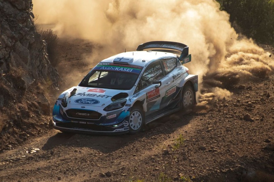 A white and blue Ford rally car speeds along a rugged dirt track, kicking up a massive cloud of thick dust behind it